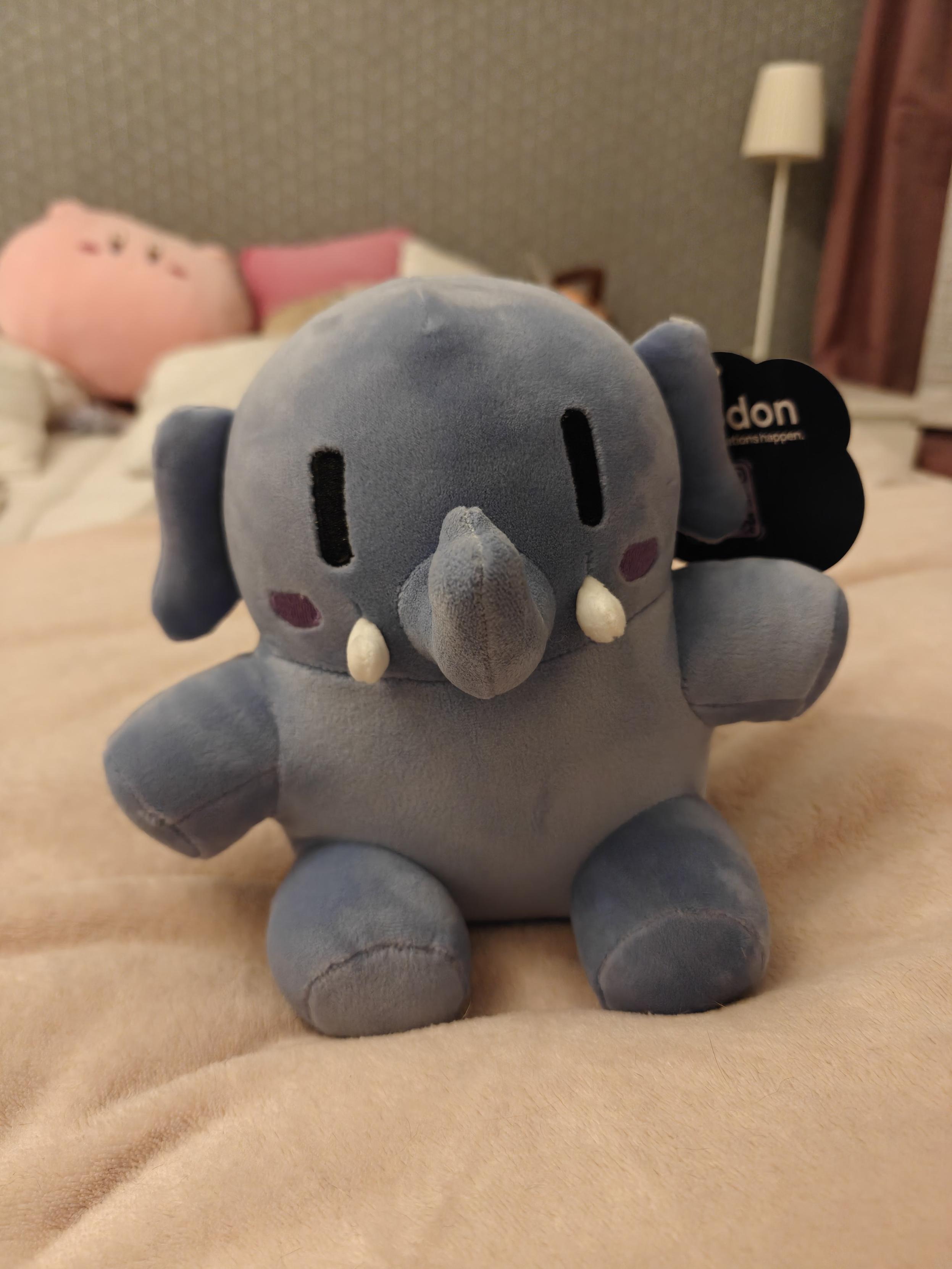 The mastodon plushie, small blue variant, sitting on a bed looking at the camera. In the background one can vaguely see other plushies, like a kirby and a pochita